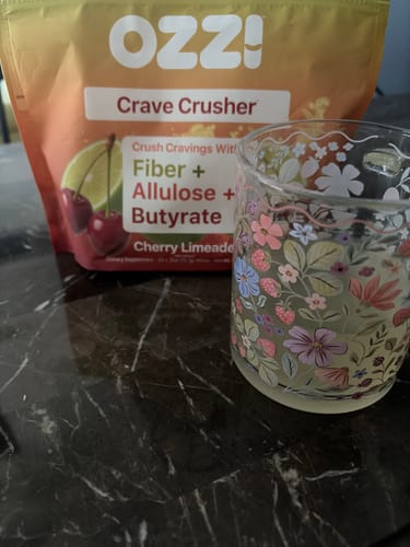 Customer's Cherry Limeade bag of Ozzi Crave Crusher behind a floral glass of the prepared drink on a marble counter.