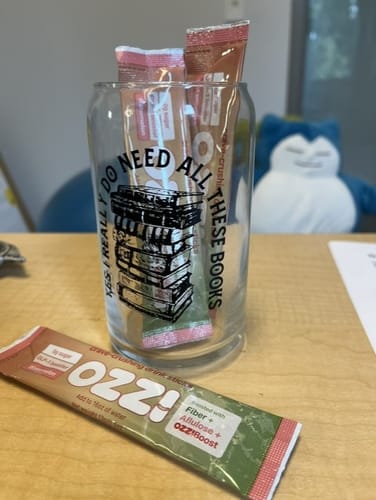 Two Ozzi drink sticks tucked into a book-themed glass on a desk, with a third stick laid in front.