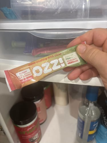 Customer holds an Ozzi crave-crushing drink stick in front of an open supplement cabinet.