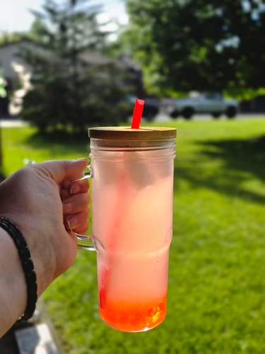 Reviewer's pink prepared Ozzi drink in a tall glass mug with a straw, held up outdoors on a sunny day.