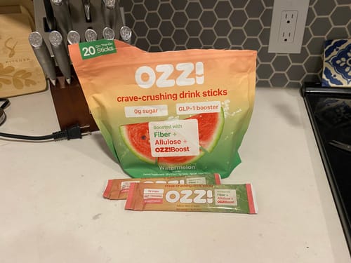 Reviewer's bag and two single sticks of Ozzi Crave Crusher drink mix in watermelon flavor, displayed on a kitchen counter.