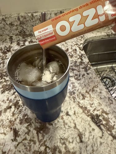 Reviewer pours a Peach Elderflower Ozzi stick into an iced tumbler on a granite counter.