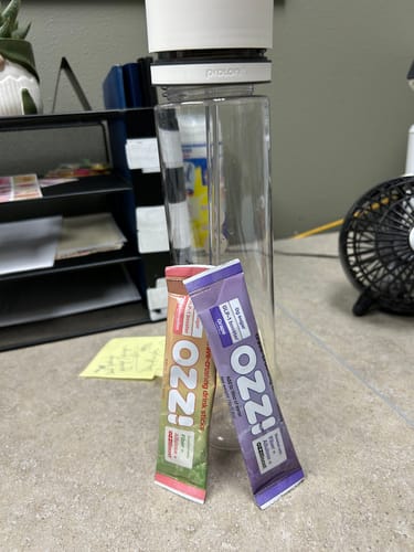 Customer displays two Ozzi drink stick packets, grape and another flavor, next to a clear water bottle on a desk.