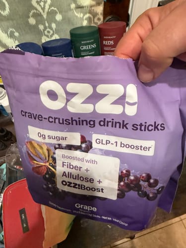 Reviewer holds a Grape bag of Ozzi drink sticks in front of a shelf of other supplements.