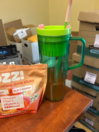 Customer's pouch of Ozzi drink sticks next to a tall green tumbler filled with the prepared beverage on a desk.